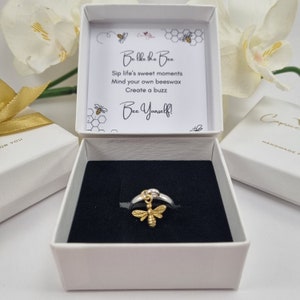 May include: A silver ring with a gold bee charm hanging from it. The ring is in a white box with a black interior. The box has a white lid with a quote about bees on it: "Be like the bee. Sip life's sweet moments. Mind your own beeswax. Create a buzz. Bee Yourself!"