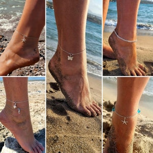 Könnte beinhalten: Vier verschiedene silberne Fußkettchen mit verschiedenen Anhängern, darunter ein Seestern, ein Schmetterling, eine Palme und eine Muschel. Die Fußkettchen werden an den Knöcheln einer Frau an einem Sandstrand gezeigt.