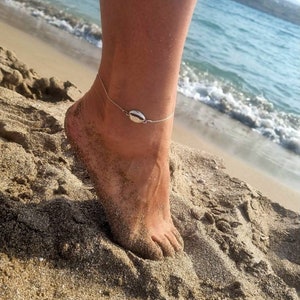 Könnte beinhalten: Ein silbernes Fußkettchen mit einem kleinen, ovalen Anhänger. Das Fußkettchen wird an einem Fußgelenk getragen, und der Hintergrund ist ein Sandstrand mit Meereswellen.