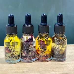 Op de afbeelding: Vier kleine glazen flesjes met zwarte druppelaars, gevuld met een heldere vloeistof en gedroogde bloemen. De flesjes zijn gelabeld met "nourishing oil", "goddess oil", "divine feminine oil" en "dream oil".