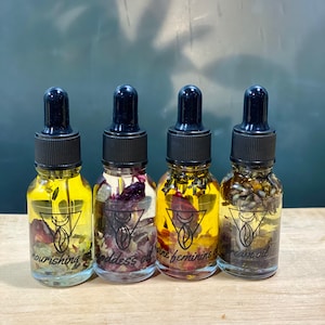 Op de afbeelding: Vier kleine glazen flesjes met druppelaars gevuld met een heldere vloeistof en gedroogde bloemen. De flesjes zijn gelabeld met "nourishing oil", "goddess oil", "divine feminine oil" en "dream oil".