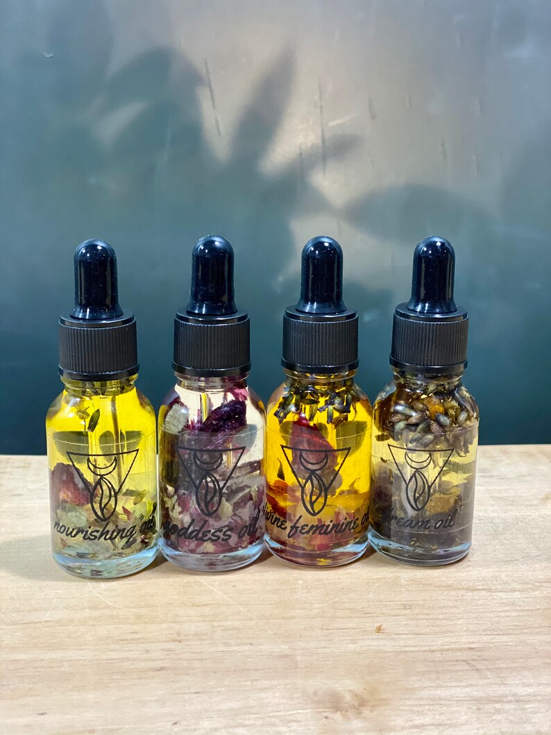 Op de afbeelding: Vier kleine glazen flesjes met druppelaars gevuld met een heldere vloeistof en gedroogde bloemen. De flesjes zijn gelabeld met "nourishing oil", "goddess oil", "divine feminine oil" en "dream oil".