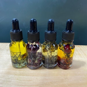 Op de afbeelding: Vier kleine glazen flesjes met druppelaars gevuld met een heldere vloeistof en verschillende gedroogde bloemen en kruiden. De flesjes zijn gelabeld met "Wishing Oil", "Goddess Oil", "Dream Oil" en "Divine Feminine Oil".