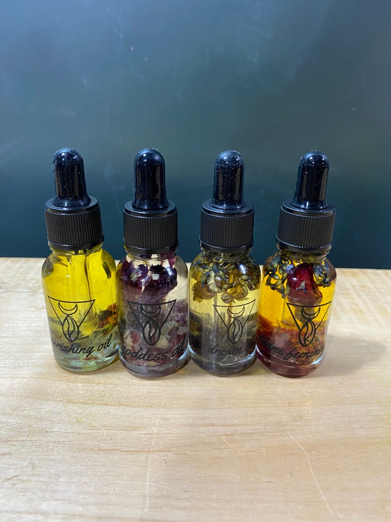 Op de afbeelding: Vier kleine glazen flesjes met druppelaars gevuld met een heldere vloeistof en verschillende gedroogde bloemen en kruiden. De flesjes zijn gelabeld met "Wishing Oil", "Goddess Oil", "Dream Oil" en "Divine Feminine Oil".