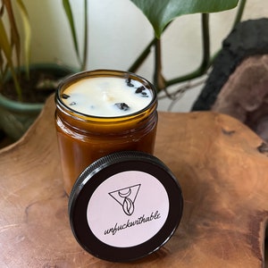 Puede incluir: Un tarro de vela de vidrio marr&oacute;n con una vela blanca y cristales negros en su interior. El tarro tiene una tapa negra con una pegatina blanca que dice "unfuckwithable" y un dise&ntilde;o de luna creciente y tri&aacute;ngulo.