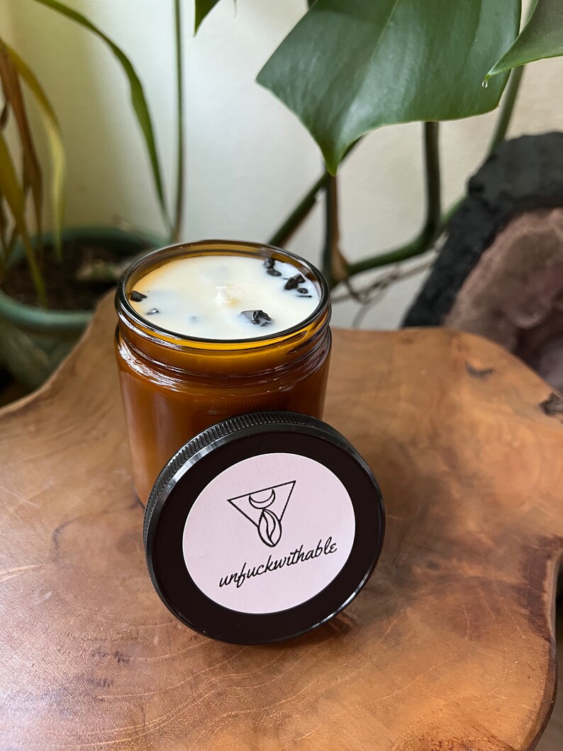 Puede incluir: Un tarro de vela de vidrio marr&oacute;n con una vela blanca y cristales negros en su interior. El tarro tiene una tapa negra con una pegatina blanca que dice "unfuckwithable" y un dise&ntilde;o de luna creciente y tri&aacute;ngulo.