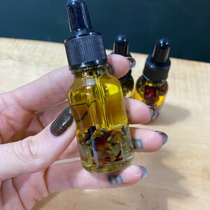 Op de afbeelding: Een glazen fles met een zwarte druppelaar, gevuld met een gele vloeistof en gedroogde bloemen en kruiden. De fles wordt in een hand met donker nagellak vastgehouden.