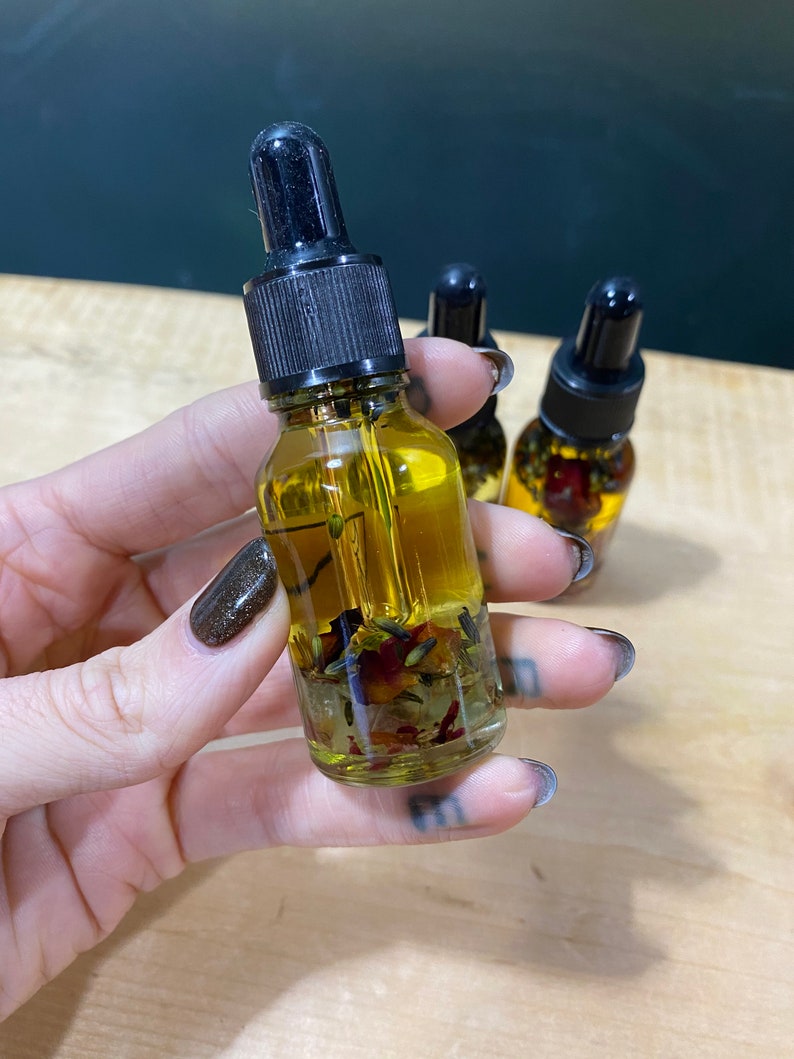 Op de afbeelding: Een glazen fles met een zwarte druppelaar, gevuld met een gele vloeistof en gedroogde bloemen en kruiden. De fles wordt in een hand met donker nagellak vastgehouden.
