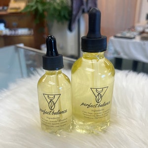 May include: Two glass bottles with dropper tops filled with a light yellow liquid. Both bottles have a black and white label with the text "perfect balance" and a list of ingredients. The ingredients include Clary Sage, Lavender, Grapefruit, Geranium EO, Sesame, Almond, Sunflower Oil, and Clear Quartz.