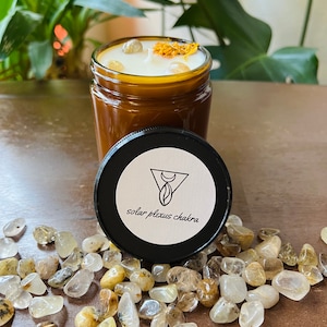 Könnte beinhalten: Eine braune Glaskerze mit einem weißen Wachsoberteil und gelben Blütenblättern. Das Glas hat einen schwarzen Deckel mit dem Text "Solarplexus-Chakra" auf einem weißen Etikett.