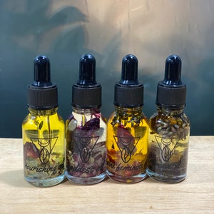 Op de afbeelding: Vier kleine glazen flesjes met druppelaars gevuld met een heldere vloeistof en gedroogde bloemen. De flesjes zijn gelabeld met "nourishing oil", "goddess oil", "wine feminine oil" en "dream oil".