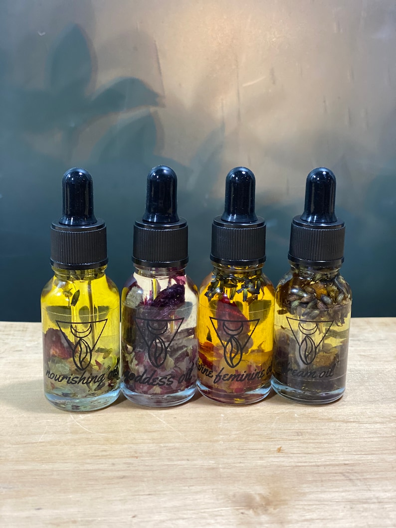 Op de afbeelding: Vier kleine glazen flesjes met druppelaars gevuld met een heldere vloeistof en gedroogde bloemen. De flesjes zijn gelabeld met "nourishing oil", "goddess oil", "wine feminine oil" en "dream oil".
