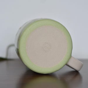May include: A handmade ceramic mug, viewed from the bottom, with a light green rim and a beige interior. The mug has a matching handle and a small, stamped design on the base. The mug is sitting on a dark brown surface.