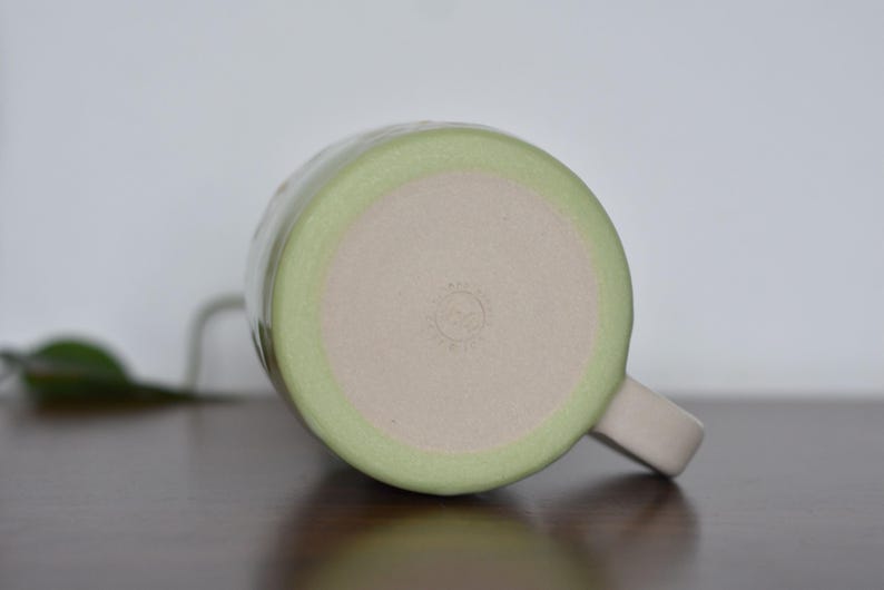 May include: A handmade ceramic mug, viewed from the bottom, with a light green rim and a beige interior. The mug has a matching handle and a small, stamped design on the base. The mug is sitting on a dark brown surface.