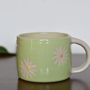 May include: A light green ceramic mug with a cream-colored handle and interior. The mug is decorated with embossed white and yellow daisy flowers. This mug is perfect for enjoying coffee or tea.
