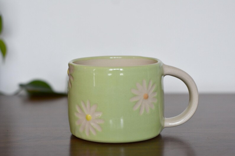 May include: A light green ceramic mug with a cream-colored handle and interior. The mug is decorated with embossed white and yellow daisy flowers. This mug is perfect for enjoying coffee or tea.