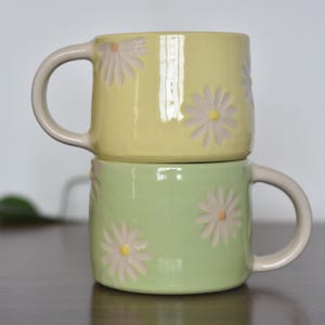 May include: Two ceramic mugs stacked on top of each other. The top mug is yellow, and the bottom mug is green. Both mugs have daisy flower designs and cream-colored handles.