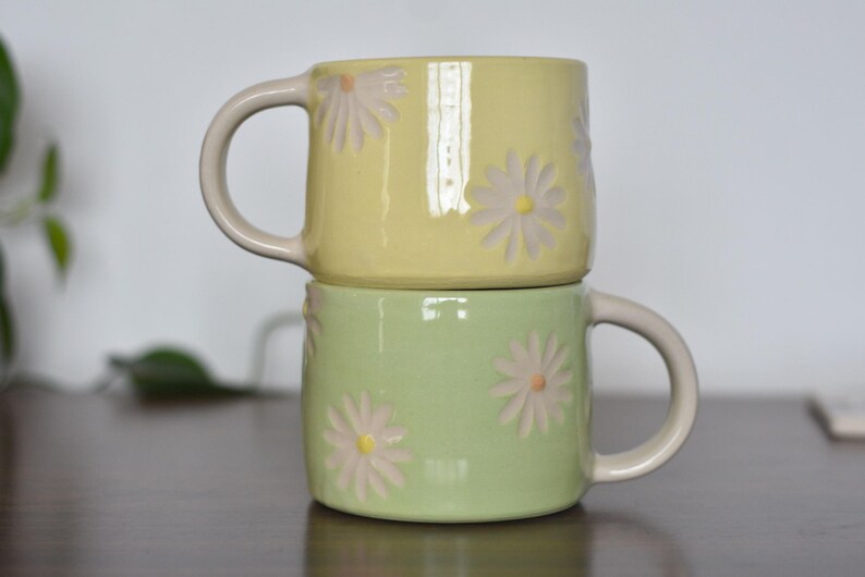 May include: Two ceramic mugs stacked on top of each other. The top mug is yellow, and the bottom mug is green. Both mugs have daisy flower designs and cream-colored handles.