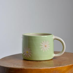 May include: A light green ceramic mug with a white interior and a handle. The mug is decorated with embossed white and pink daisy flowers. The mug is sitting on a small wooden stool.