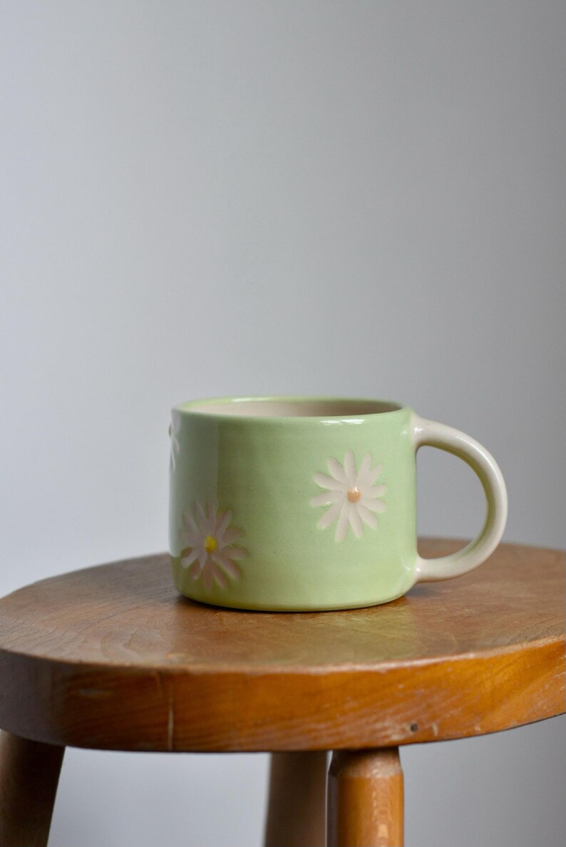 May include: A light green ceramic mug with a white interior and a handle. The mug is decorated with embossed white and pink daisy flowers. The mug is sitting on a small wooden stool.