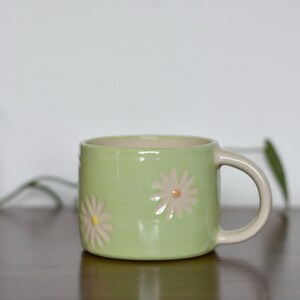 May include: A light green ceramic mug with a beige handle and interior. The mug features embossed daisy designs in a light pink color. The mug is sitting on a dark brown surface.