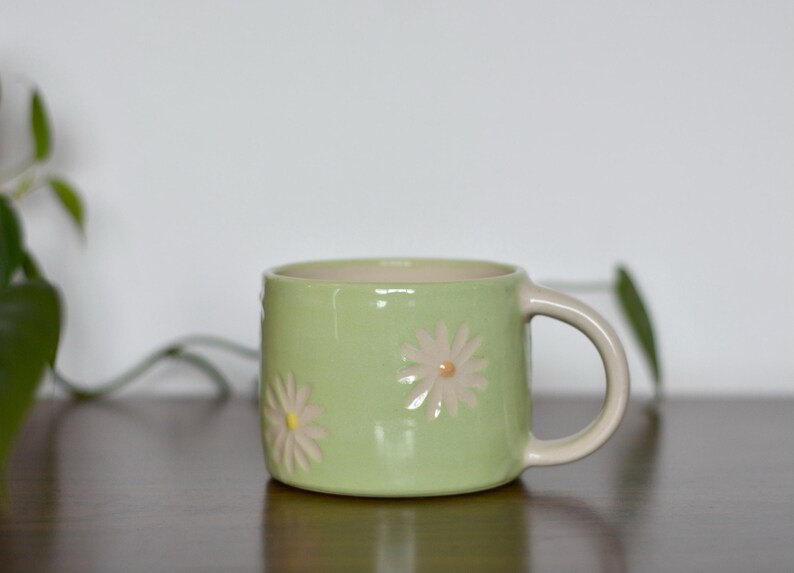 May include: A light green ceramic mug with a beige handle and interior. The mug features embossed daisy designs in a light pink color. The mug is sitting on a dark brown surface.