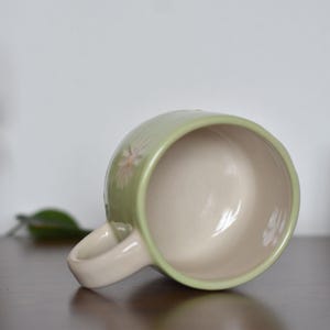 May include: A light green ceramic mug with a cream-colored interior and handle. The mug features a floral design and is resting on its side. Another blue mug is visible in the background.