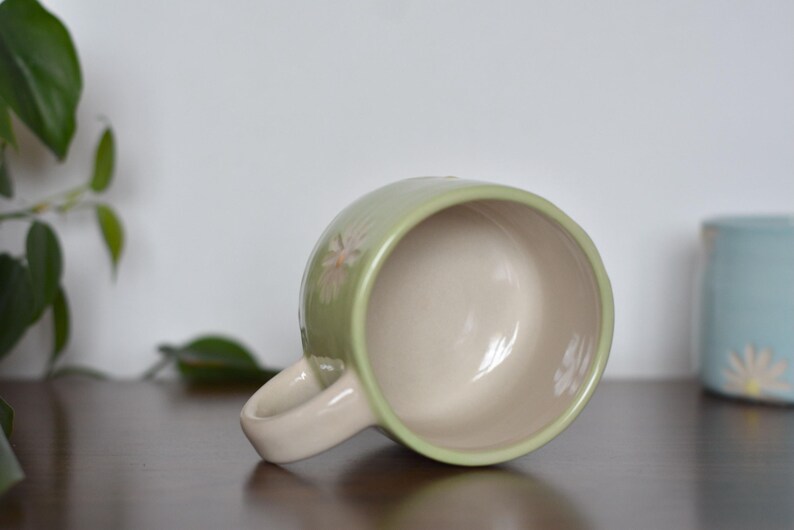 May include: A light green ceramic mug with a cream-colored interior and handle. The mug features a floral design and is resting on its side. Another blue mug is visible in the background.