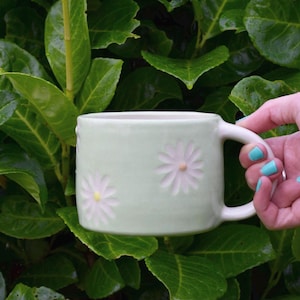 May include: A light green ceramic mug with a white handle and embossed daisy flowers. The mug is held by a hand with turquoise nail polish against a backdrop of green leaves. The mug is approximately 4 inches tall.