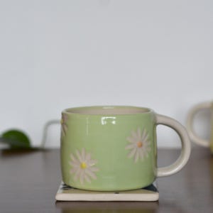 May include: A light green ceramic mug with daisy flower designs. The mug has a cream-colored handle and is sitting on a small square coaster. A second yellow mug with daisy designs is in the background.