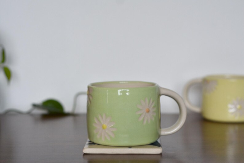 May include: A light green ceramic mug with daisy flower designs. The mug has a cream-colored handle and is sitting on a small square coaster. A second yellow mug with daisy designs is in the background.
