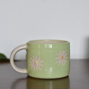 May include: A light green ceramic mug with a cream-colored handle and interior. The mug features embossed daisy designs with yellow centers. Another blue mug with a similar design is in the background.