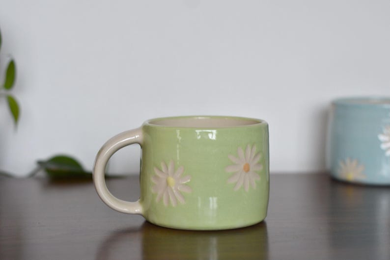 May include: A light green ceramic mug with a cream-colored handle and interior. The mug features embossed daisy designs with yellow centers. Another blue mug with a similar design is in the background.