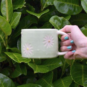 May include: A light green ceramic mug with two daisy designs. The mug is held by a hand with blue nail polish against a backdrop of green leaves. The mug has a square shape and a white handle.