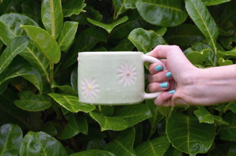 May include: A light green ceramic mug with two daisy designs. The mug is held by a hand with blue nail polish against a backdrop of green leaves. The mug has a square shape and a white handle.