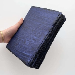 Op de afbeelding: Een stapel handgemaakt papier met een donkerblauw gemarmerd ontwerp. De randen van het papier zijn ruw en ongelijkmatig. Het papier wordt vastgehouden in een hand met een gouden armband.