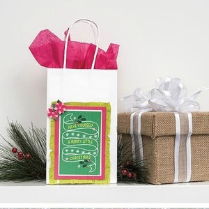 Pu&ograve; includere: Una borsa regalo bianca con una carta di Natale verde e rossa attaccata. La carta ha uno striscione verde con il testo "Have yourself a merry little Christmas". La borsa &egrave; piena di carta velina rosa e si trova su un tavolo accanto a una scatola regalo marrone con un nastro bianco.