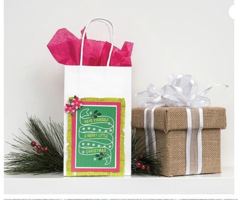 Pu&ograve; includere: Una borsa regalo bianca con una carta di Natale verde e rossa attaccata. La carta ha uno striscione verde con il testo "Have yourself a merry little Christmas". La borsa &egrave; piena di carta velina rosa e si trova su un tavolo accanto a una scatola regalo marrone con un nastro bianco.