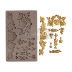 May include: A silicone mold with a variety of decorative designs, including roses, leaves, and seashells. The mold is gray and has the text "tre-design" printed on it. The mold is shown next to a set of gold-colored decorative pieces that have been made using the mold.