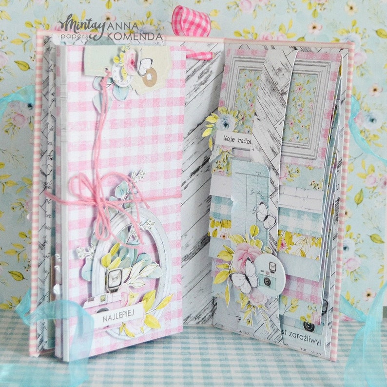 May include: A handmade scrapbook with pink gingham and floral patterned pages. The open book features decorative elements like a white frame, paper flowers, butterflies, and the word "NAJLEPIEJ". The overall theme is a vintage, feminine aesthetic.