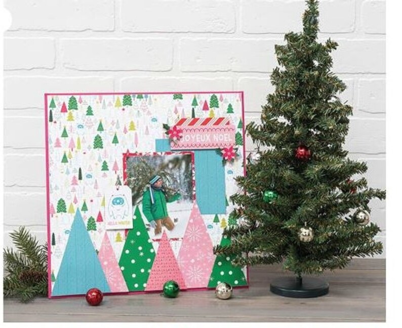 Pu&ograve; includere: Una pagina di scrapbooking rosa e blu con una foto di un bambino che indossa una giacca rosa e blu. La pagina &egrave; decorata con alberi di carta, fiocchi di neve e uno striscione che dice "Joyeux Noel".