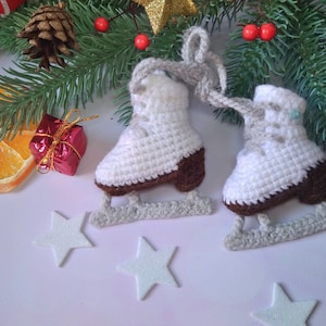 May include: Two white crocheted ice skate ornaments with brown soles and gray metal blades. The ornaments are decorated with white and gray yarn and are hanging from a green pine branch with red berries.