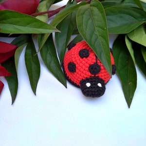 Peut inclure: Une coccinelle en crochet rouge et noire avec des points noirs et des yeux blancs. La coccinelle est posée sur une surface blanche avec des feuilles vertes.