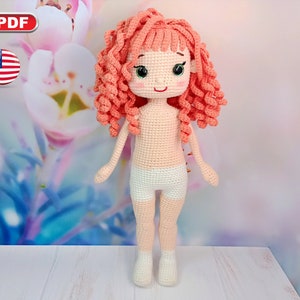 May include: Crocheted doll with pink curly hair, green eyes, and a white crocheted outfit. The doll is standing on a white wooden surface.