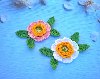 May include: Two crocheted flowers with green leaves on a blue background. One flower is pink with an orange center, and the other is white with a yellow center.