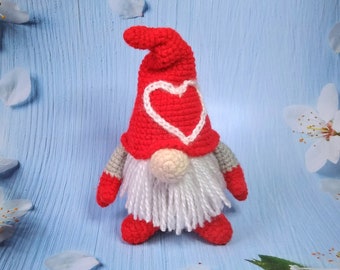May include: A crocheted gnome with a red hat and white beard. The hat has a white heart design on the front. The gnome is standing on a blue background.