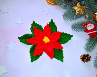 May include: A red and green crocheted poinsettia flower with a yellow center. The flower is on a white background with white stars and a pine branch with gold ornaments.