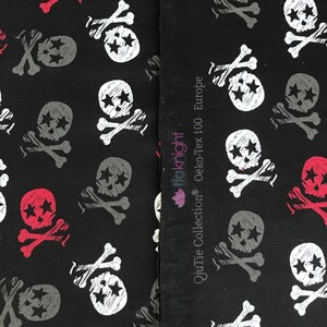 May include: Black fabric with a repeating pattern of white and gray skull and crossbones with three stars on each skull. The fabric has a label that reads "Oeko-Tex 100 Europe" and "Qutie Collection&reg;" with a pink flower logo.