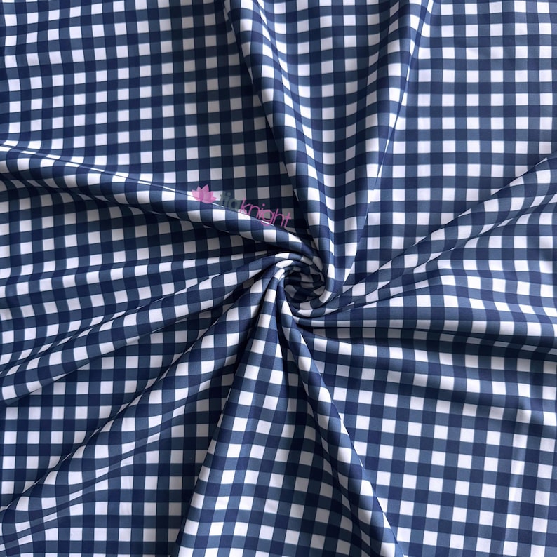K&ouml;nnte beinhalten: Ein Stoff mit einem klassischen marineblauen und wei&szlig;en Vichy-Karo-Muster. Das Material ist in der Mitte gerafft, wodurch weiche Falten und Drapierungen entstehen. Ein kleines rosa Logo mit dem Text "itaknight" ist auf der Stoffoberfl&auml;che sichtbar.