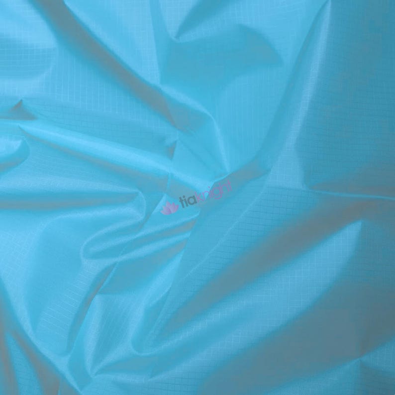 May include: A close-up of a light blue fabric with a subtle grid pattern. The fabric is folded and draped, creating soft shadows and highlights. The overall texture appears smooth and slightly shiny.